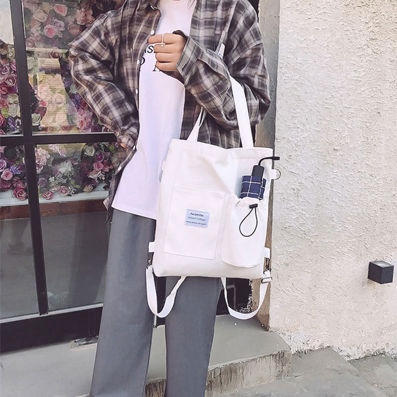 Person holding a white tote bag with a small blue tag, standing against a light-colored wall.