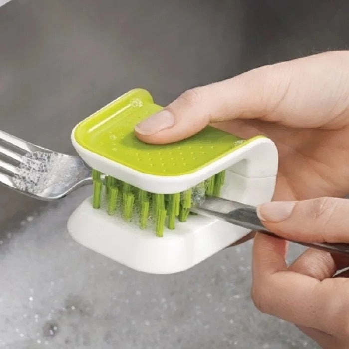 Hand using a green and white scrubber to clean a fork over a sink.