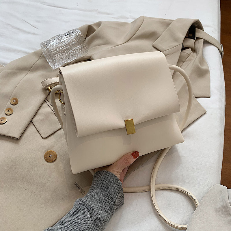 Beige handbag held by a person with a beige coat and glass on a white surface