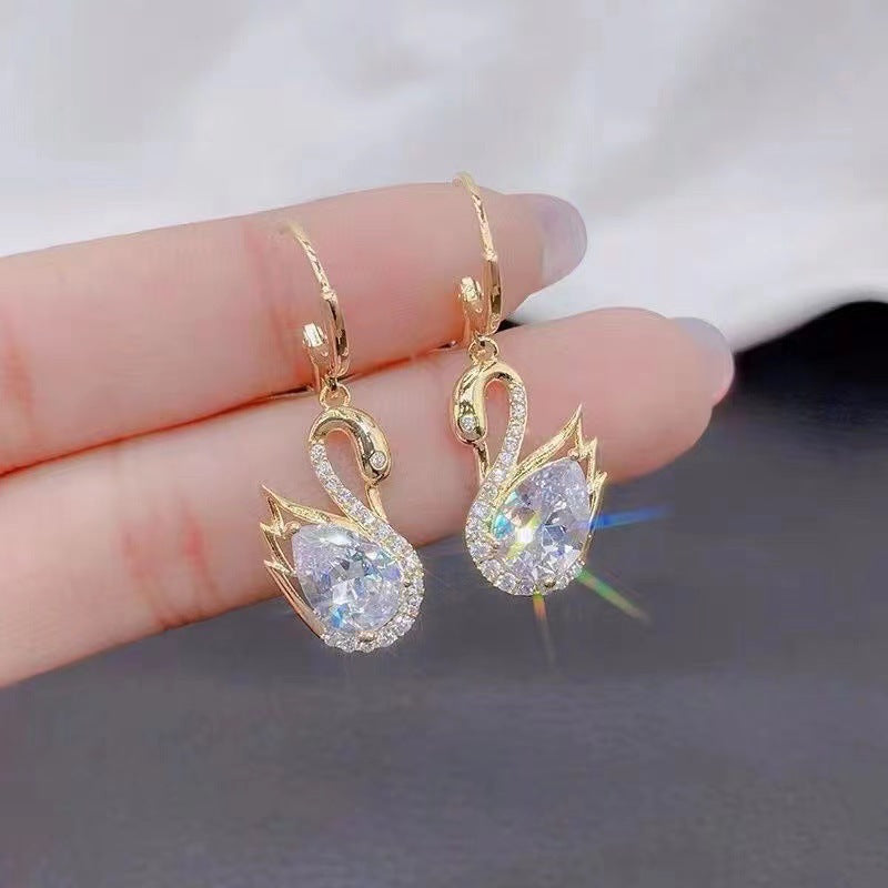 Gold Swan-shaped earrings on a finger against a neutral background