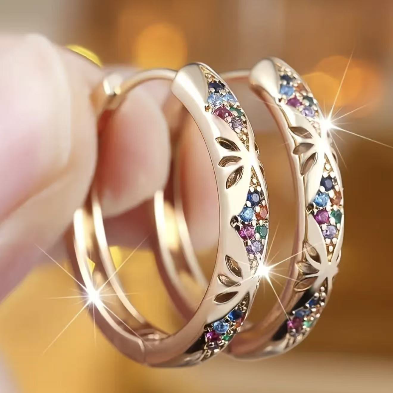 Gold hoop earrings with colorful gemstones held by a hand against a blurred background