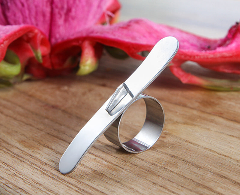 Silver ring with a flat top and curved band on a wooden surface with pink flowers in the background.