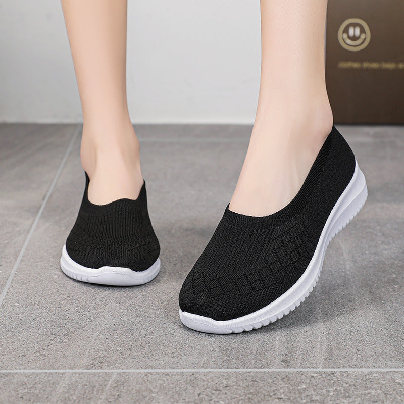 Black slip-on shoes with white soles worn by a person on a gray floor.