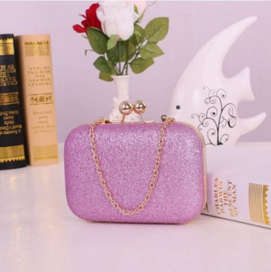 Pink glittery clutch bag with a chain strap on a surface with books and a vase in the background.