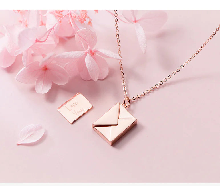 Rose gold envelope pendant necklace on a pink background with flowers