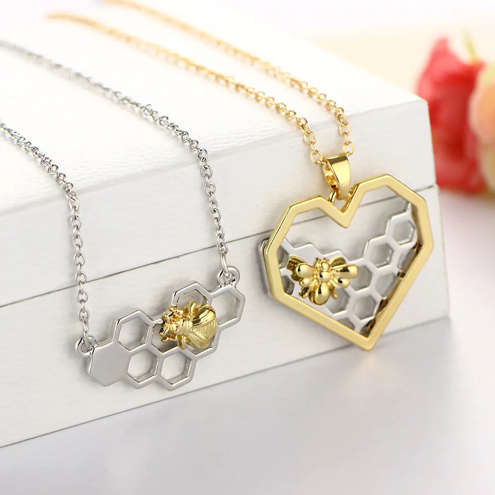 Two honeycomb-shaped necklaces, one gold and one silver, with bee details on a white surface.