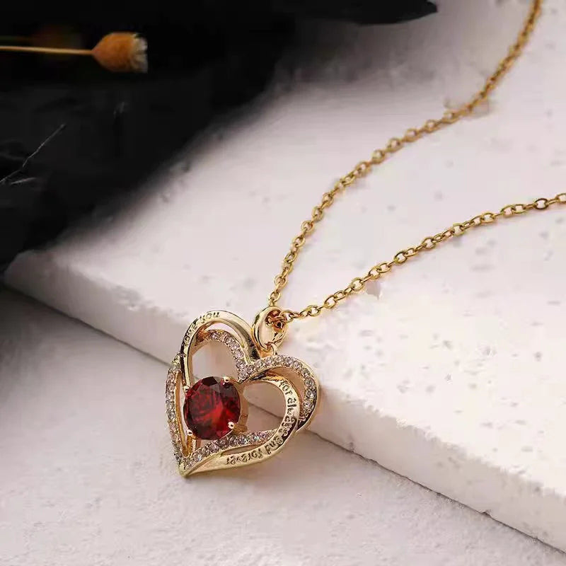 Gold necklace with a heart-shaped pendant featuring a red gemstone on a light surface.