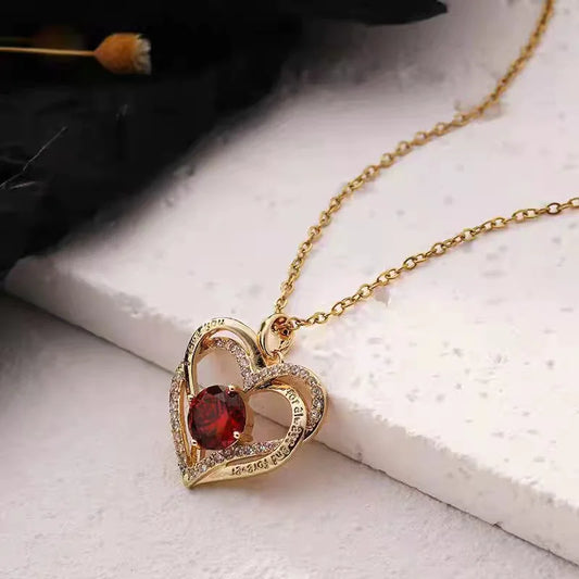 Gold necklace with a heart-shaped pendant featuring a red gemstone on a light surface.