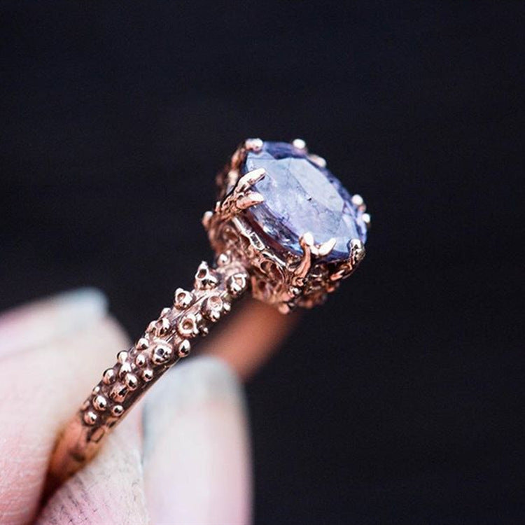 Rose gold ring with a blue gemstone held by a hand against a dark background