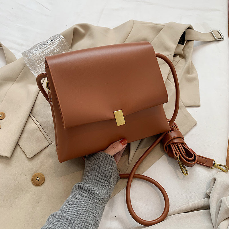 Brown leather handbag with gold clasp held by a person wearing gray sleeves, on a beige fabric background.