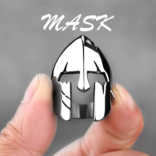 Metallic mask-shaped object held between fingers with 'MASK' text above on a gray background