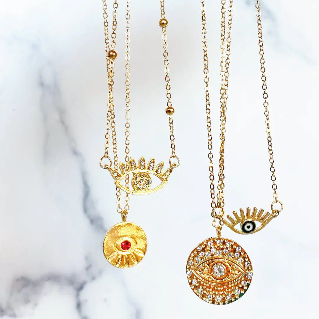 Two gold necklaces with eye-shaped pendants on a marble background