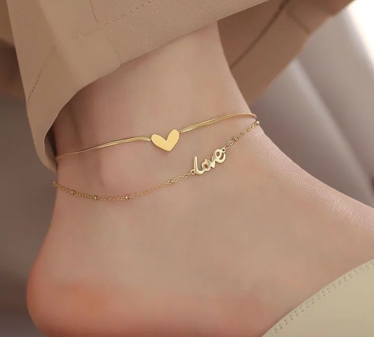 Gold anklets with heart and 'love' design on a foot against a neutral background