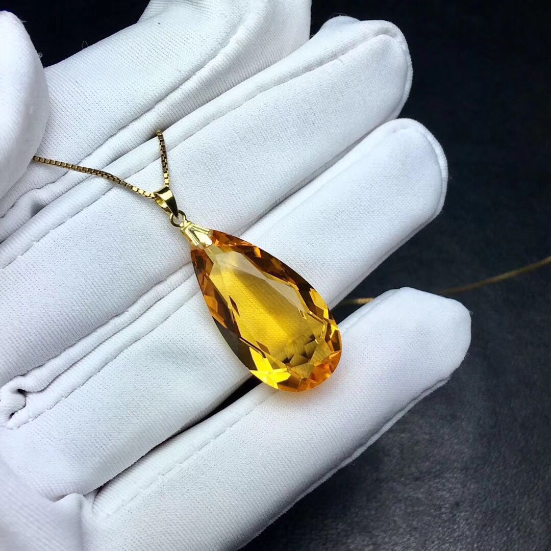 Yellow teardrop pendant on a gold chain held by a white gloved hand against a dark background