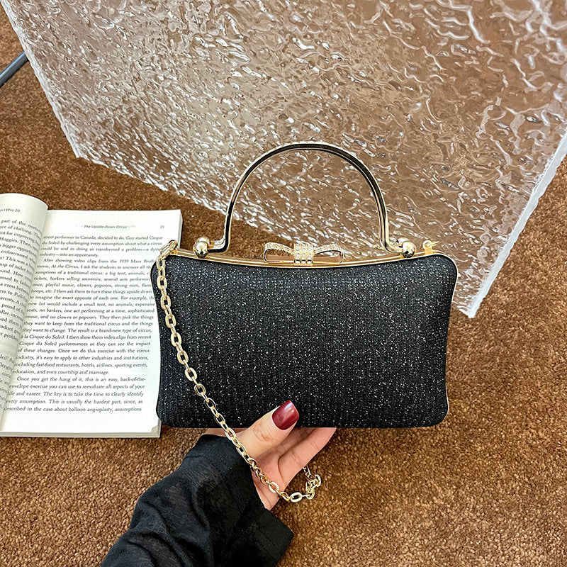 Black glittery handbag with gold chain held by a hand next to an open book on a textured surface.