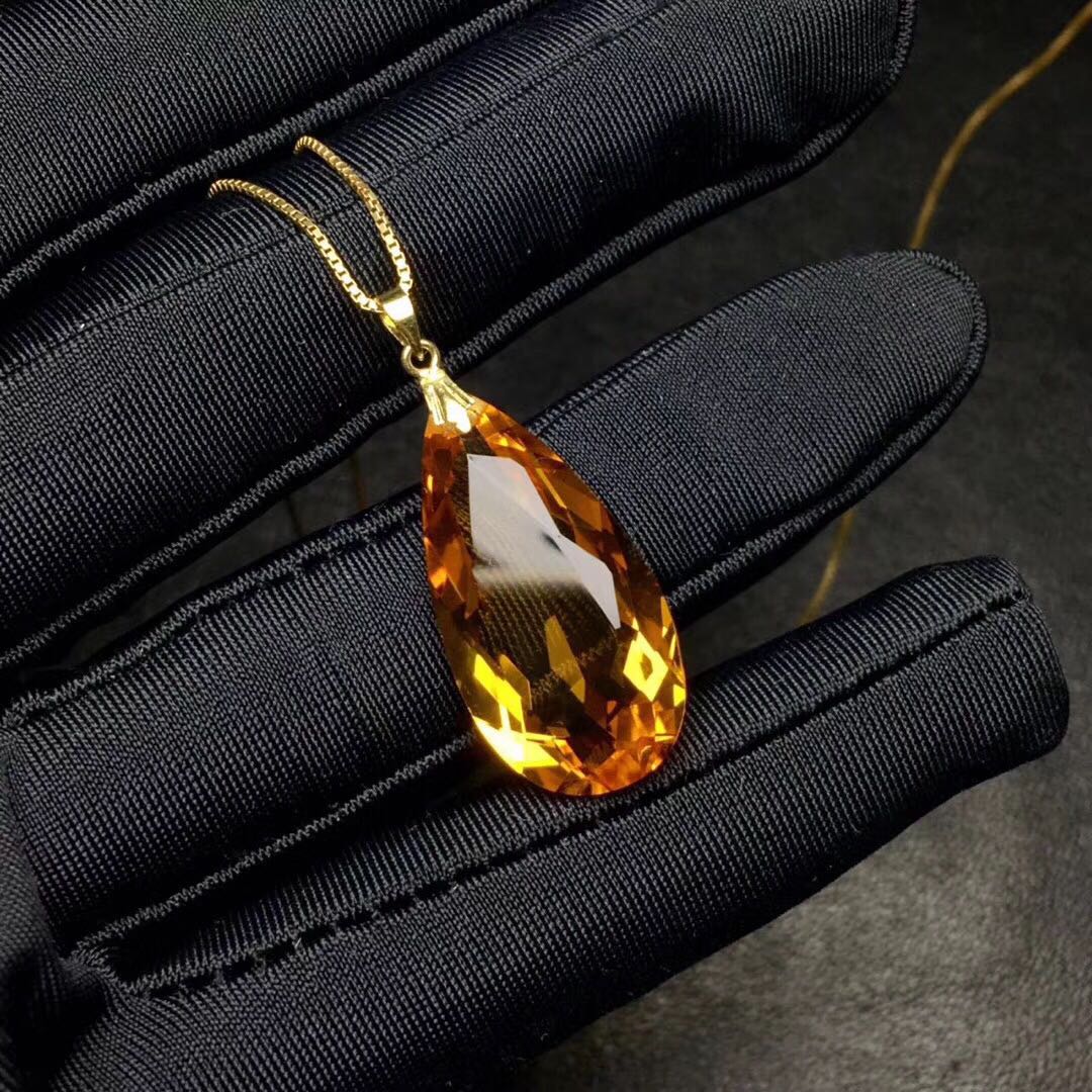 Teardrop-shaped amber pendant on a gold chain held by black gloves against a dark background