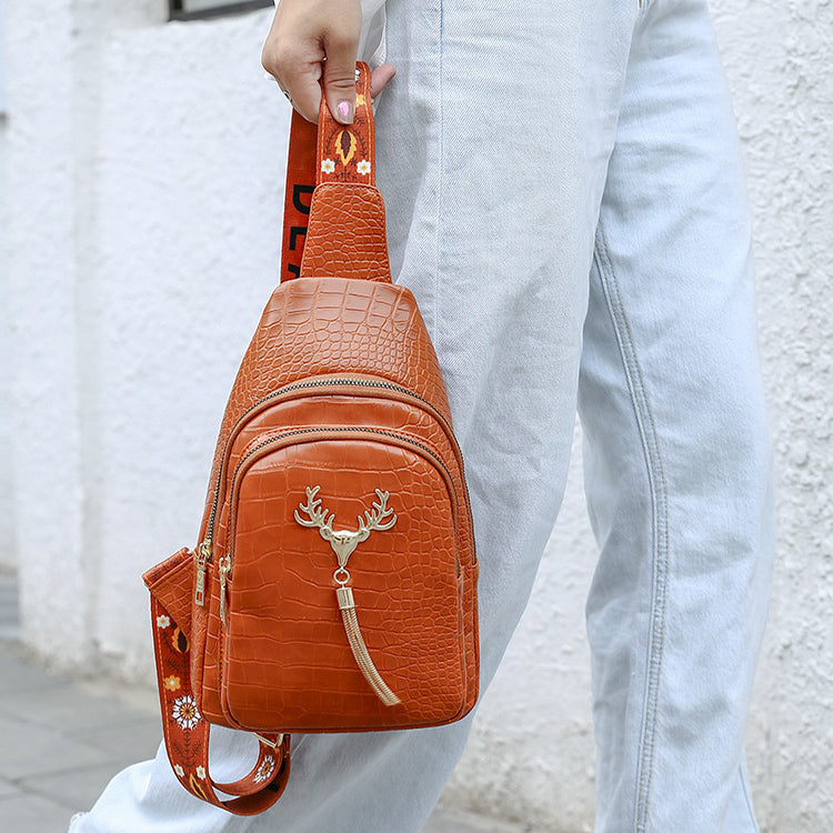 Brown crocodile-patterned sling bag with a deer emblem held by a person wearing light blue jeans.
