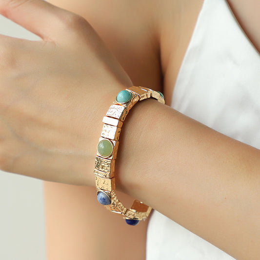 Gold bracelet with green and blue stones on a wrist against a neutral background