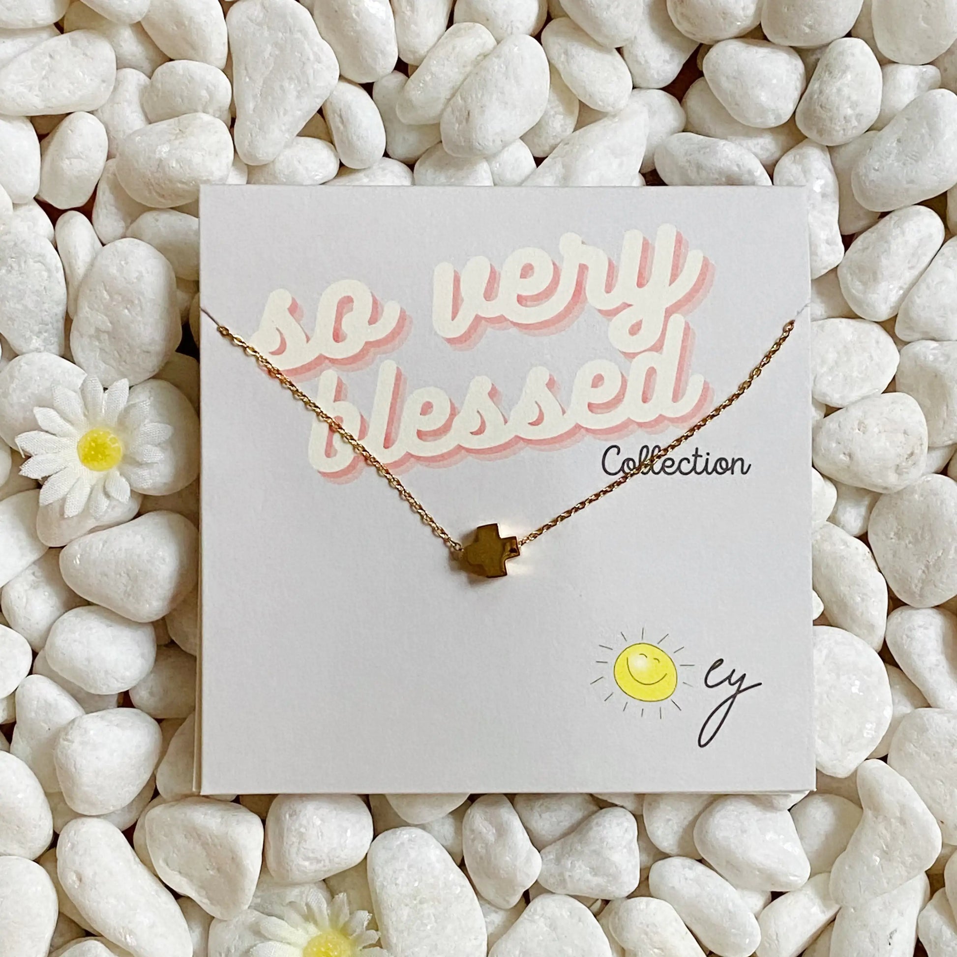 Gold necklace on a card with 'so very blessed' text, placed on white pebbles.