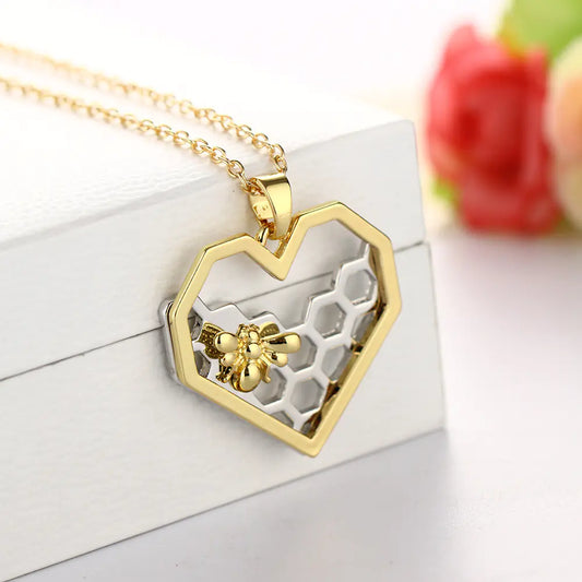 Gold necklace with a heart-shaped pendant featuring a honeycomb and bee design on a white surface.