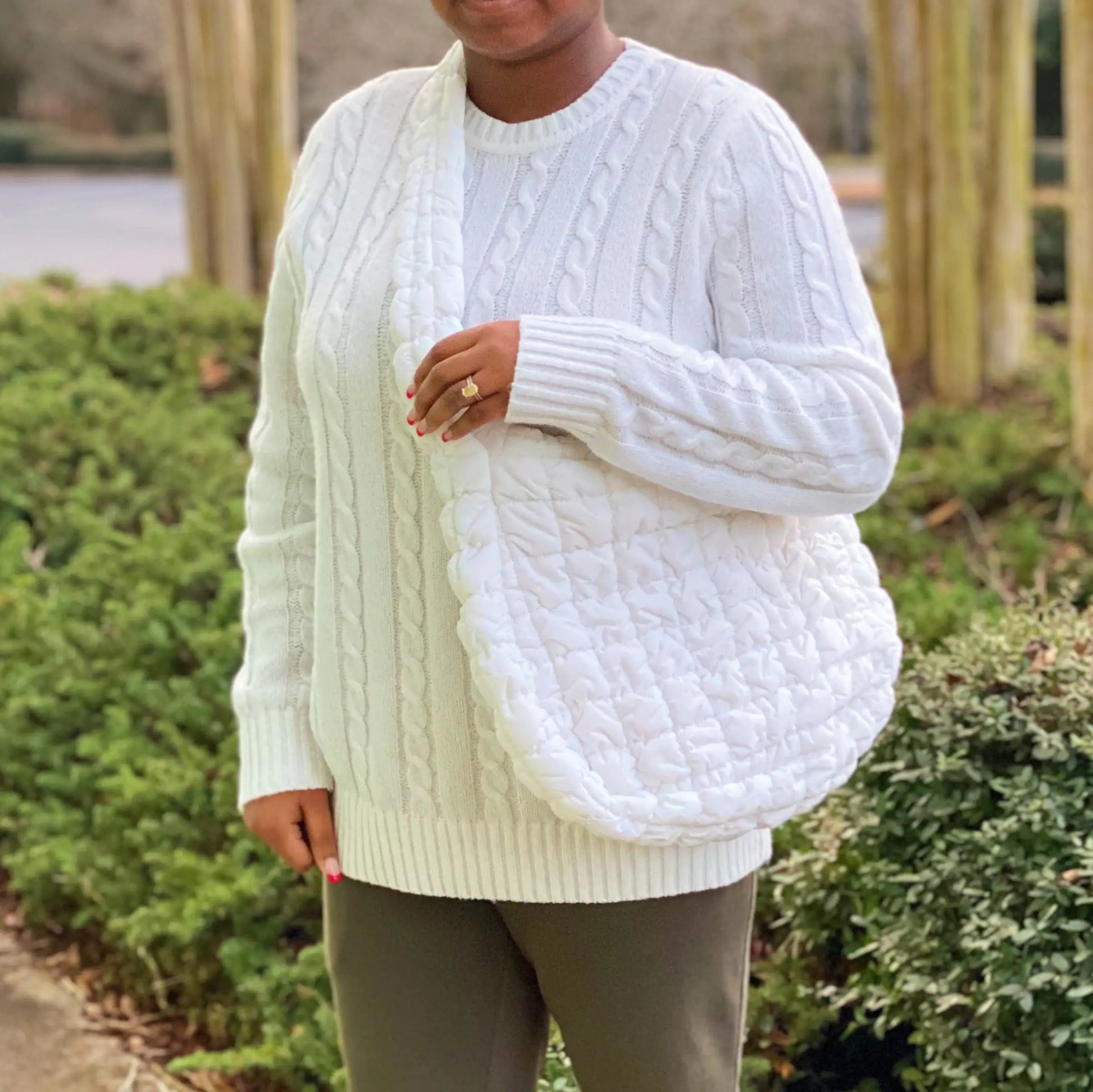 Person wearing a white cable knit sweater with a textured white bag outdoors.